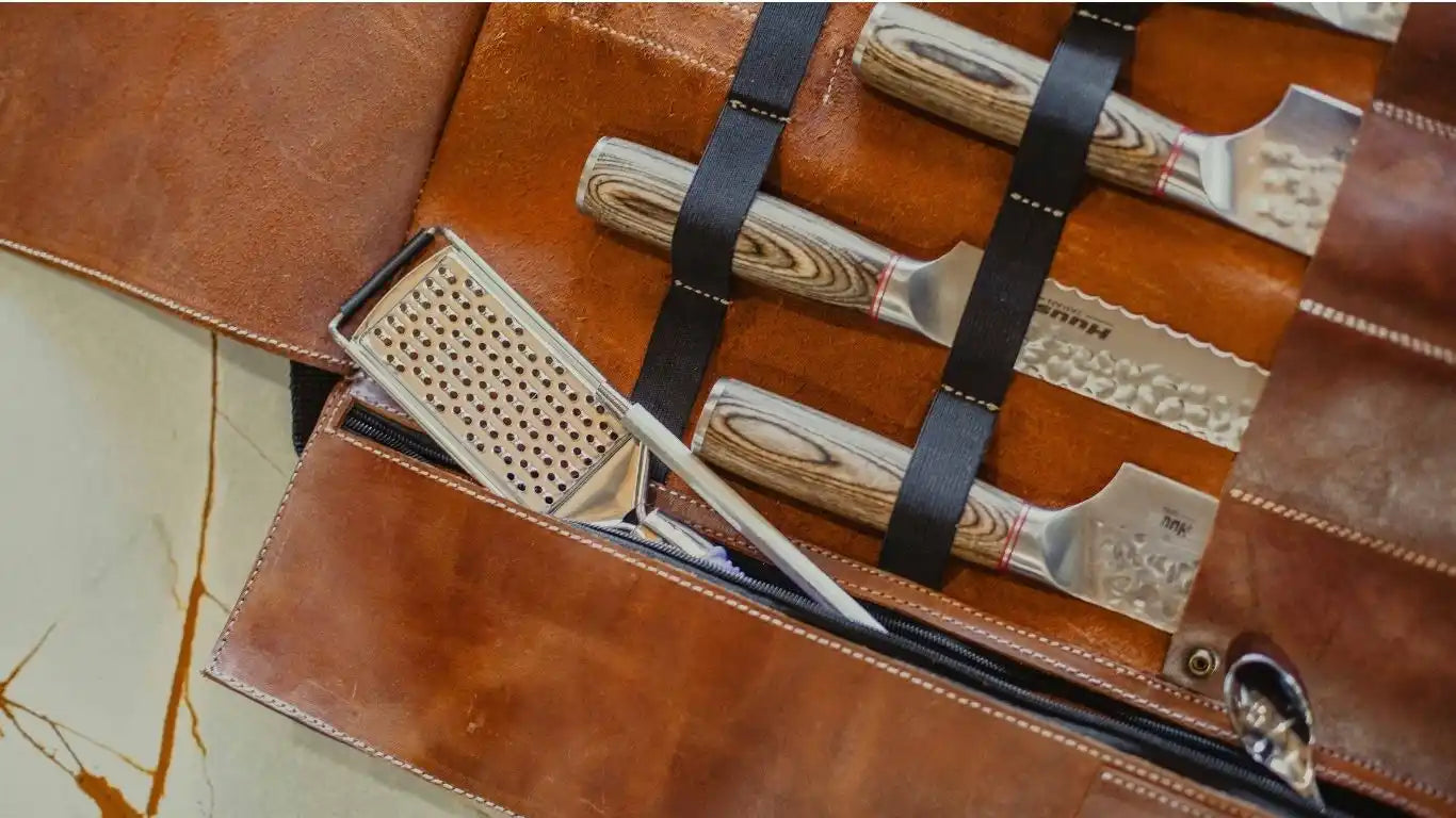 A brown leather knife roll containing several knives with patterned blades and light wood handles, and a small grater.