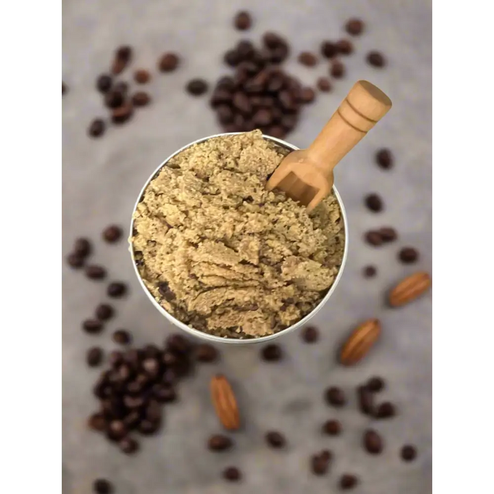 A metallic measuring cup with coarse brown coffee grounds and wooden scoop for organic coconut coffee scrubs