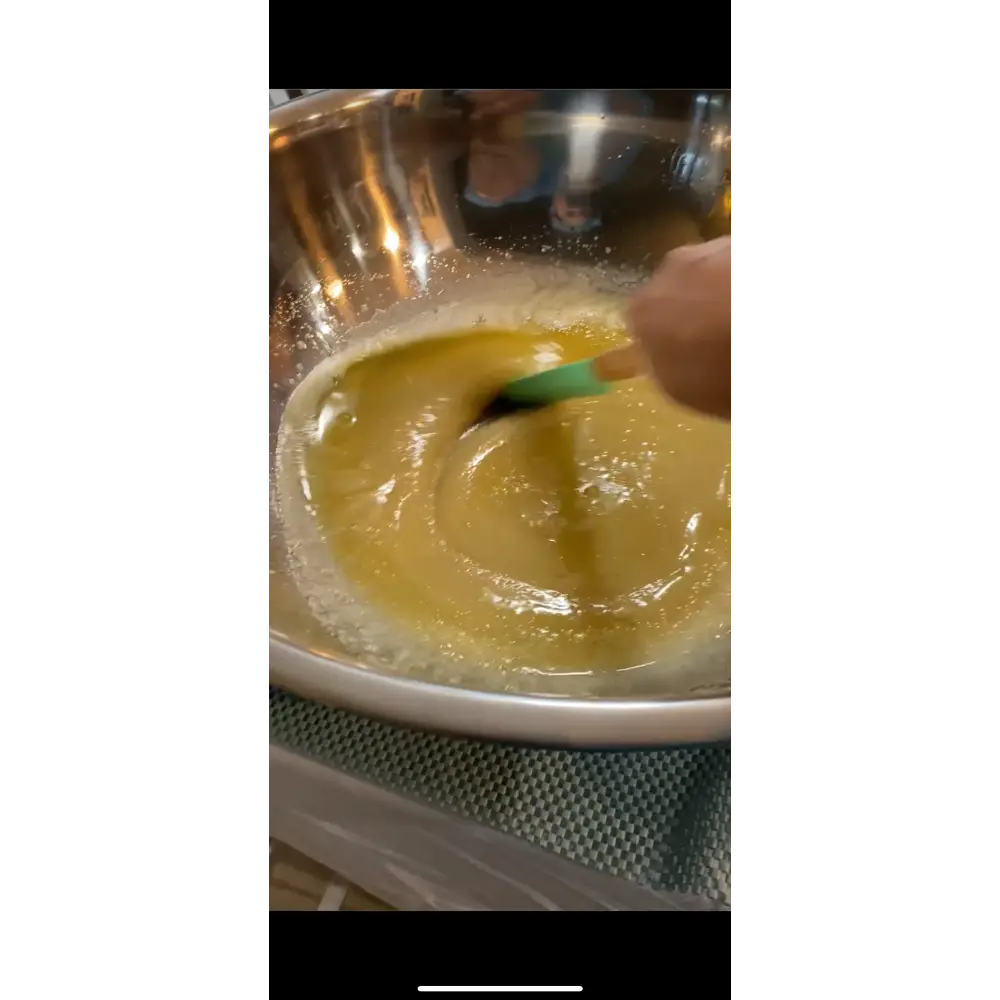 A hand stirs thick golden-yellow batter with a green silicone spatula in a stainless bowl for wendala’s crab lemon clove using pure cane sugar