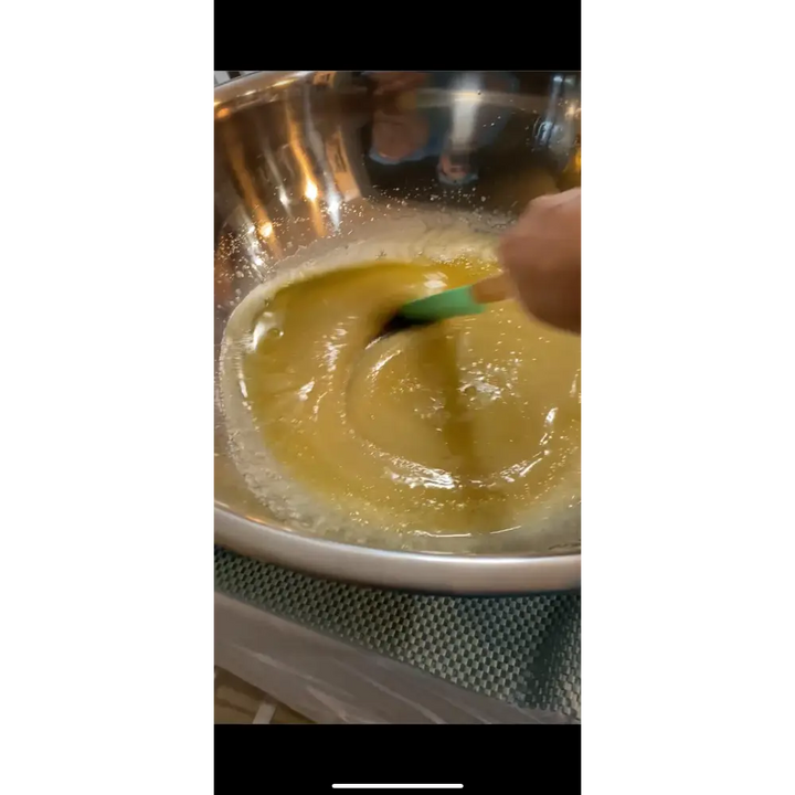 A hand stirs thick golden-yellow batter with a green silicone spatula in a stainless bowl for wendala’s crab lemon clove using pure cane sugar