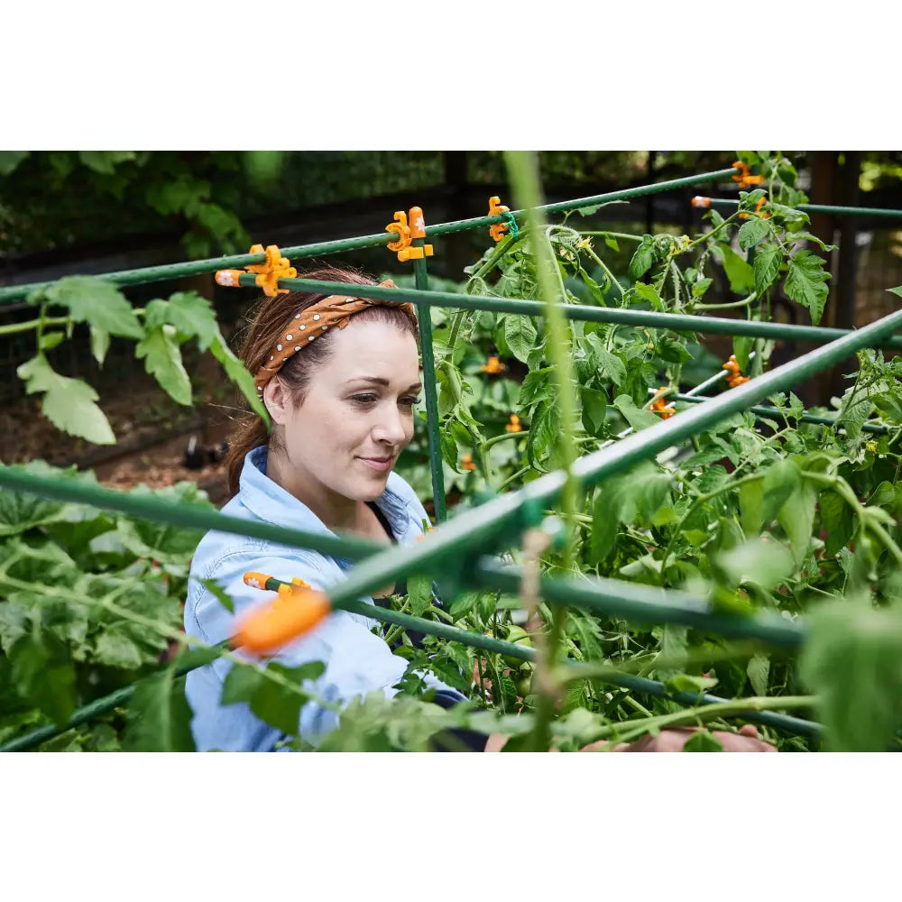 Woman in light blue denim jacket with orange headband stands among green tomato plants supported by custom trellis monster garden plant tomato cage
