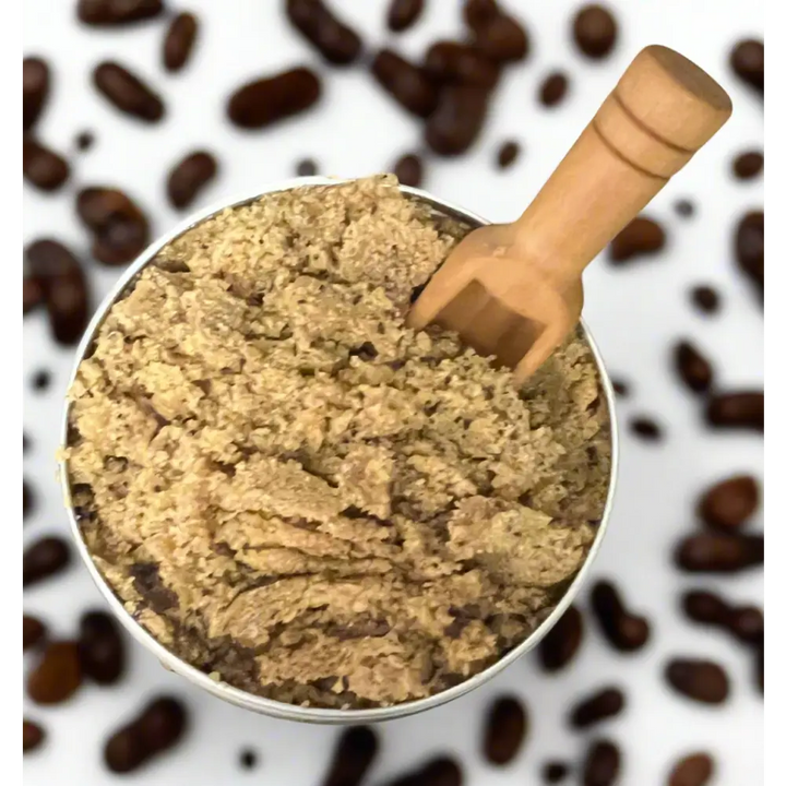 A metallic container of coarse light brown coffee scrubs with brown sugar and a wooden scoop for exfoliating feet
