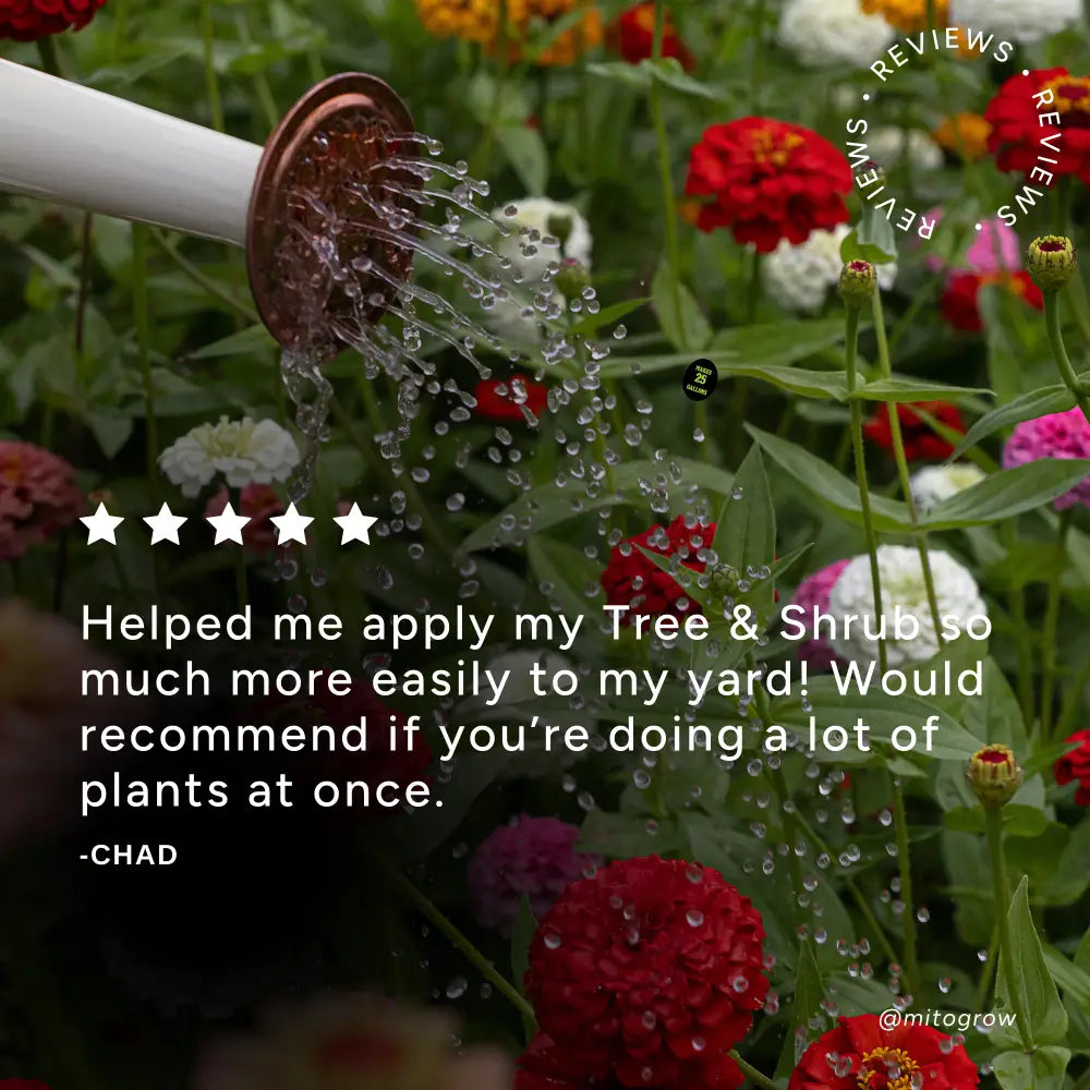 A white garden sprayer hose with a copper nozzle spraying water over colorful flowers in a garden setting