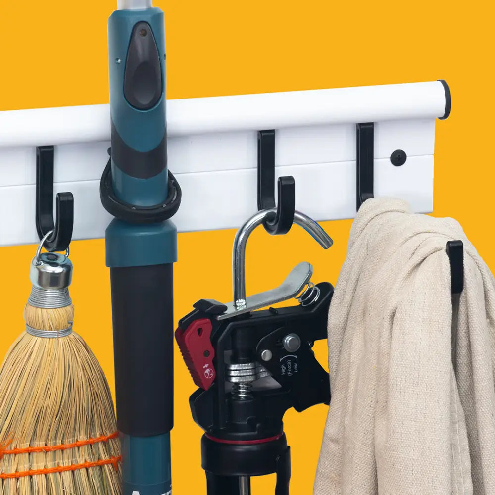 White wall-mounted universal storage hanger with black rubber hooks holding cleaning tools and a beige towel storage rack