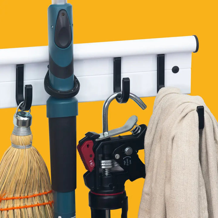White wall-mounted universal storage hanger with black rubber hooks holding cleaning tools and a beige towel storage rack