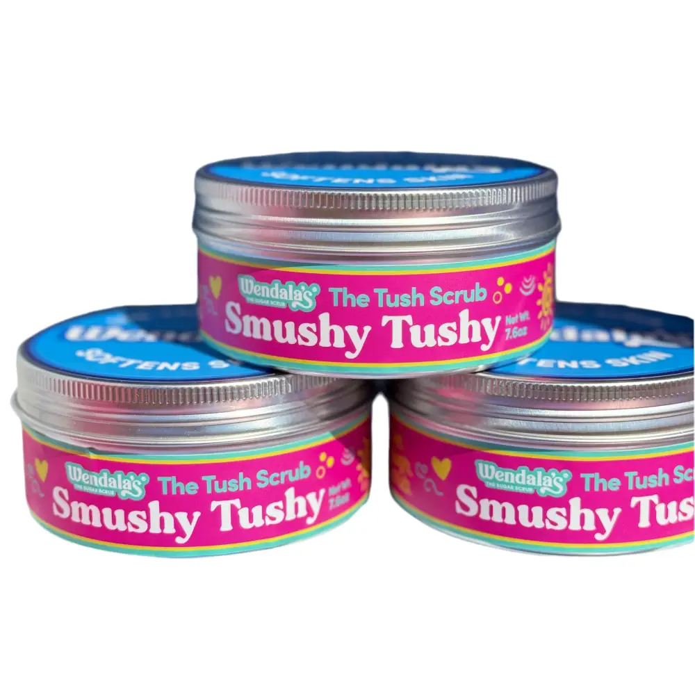 Pink and blue labeled metal jars of wendalas the tush scrub smushy tushy body scrub featuring organic coconut scent