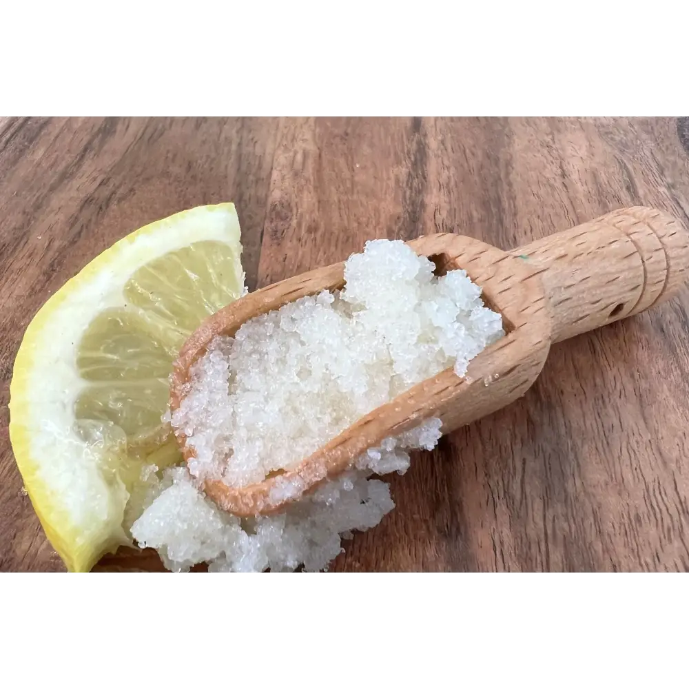 A wooden scoop of white crystalline hand scrub with organic coconut beside a lemon slice on rustic wood surface
