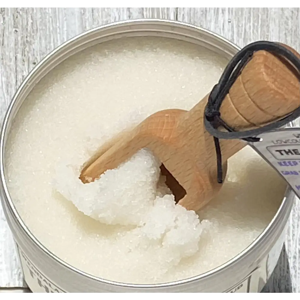 A wooden spoon with a black cord handle rests in a silver tin of lemon clove hand scrub with essential oil sugar scrub