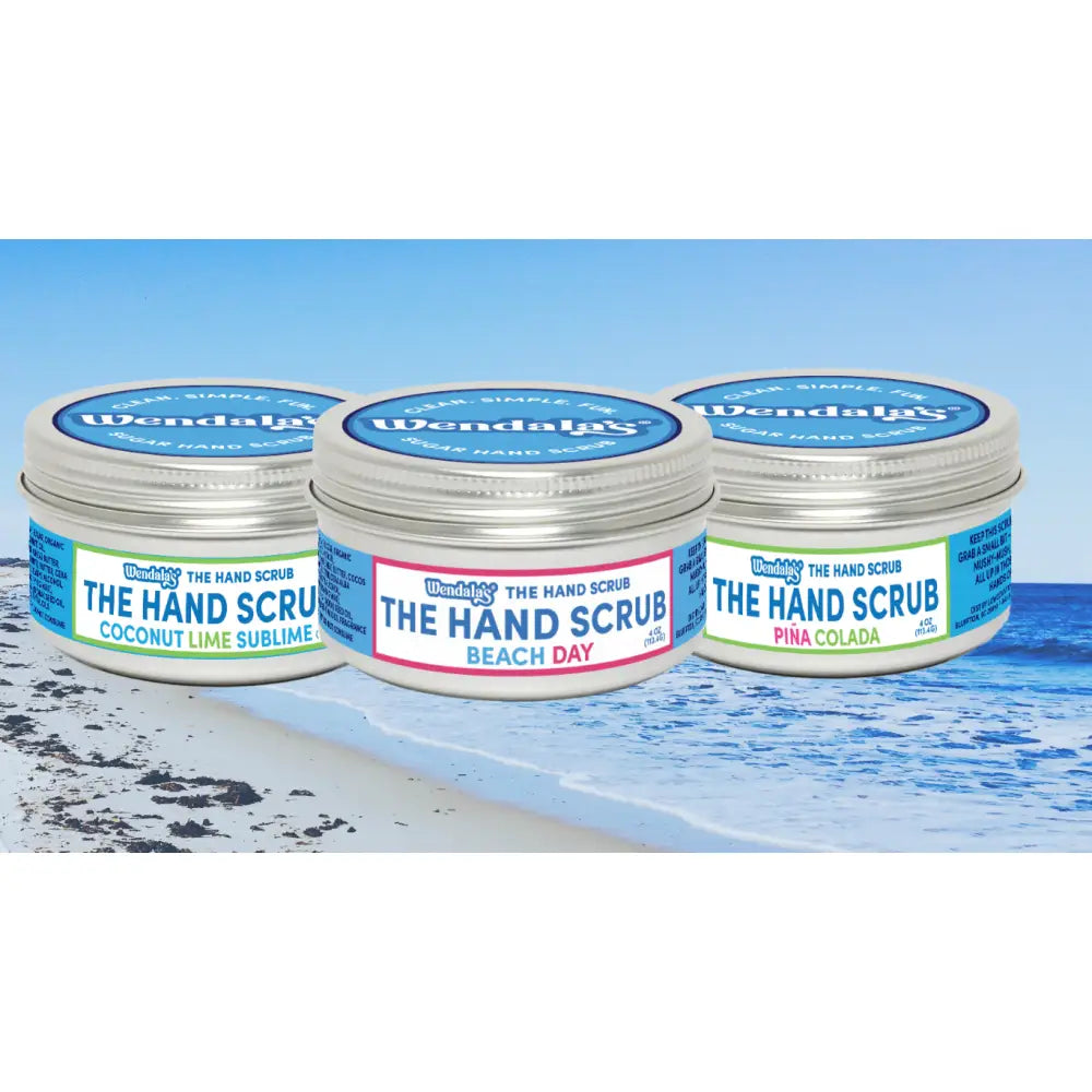 Three cylindrical metal jars of wendala’s®️ coastal hand scrub in coconut lime sublime green beach day pink and piña colada blue