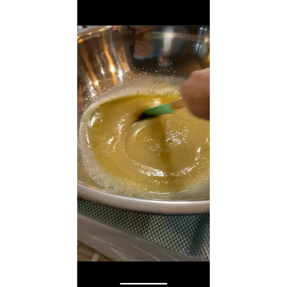 A hand stirs thick golden-yellow lemon clove batter with a green spatula in a stainless bowl for wendala’s crab organic coconut oil scrub