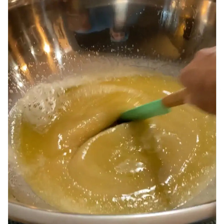 A green silicone spatula with wooden handle stirring a yellow foamy mixture in a bowl for wendala’s crab rosemary mint sugar hand scrub