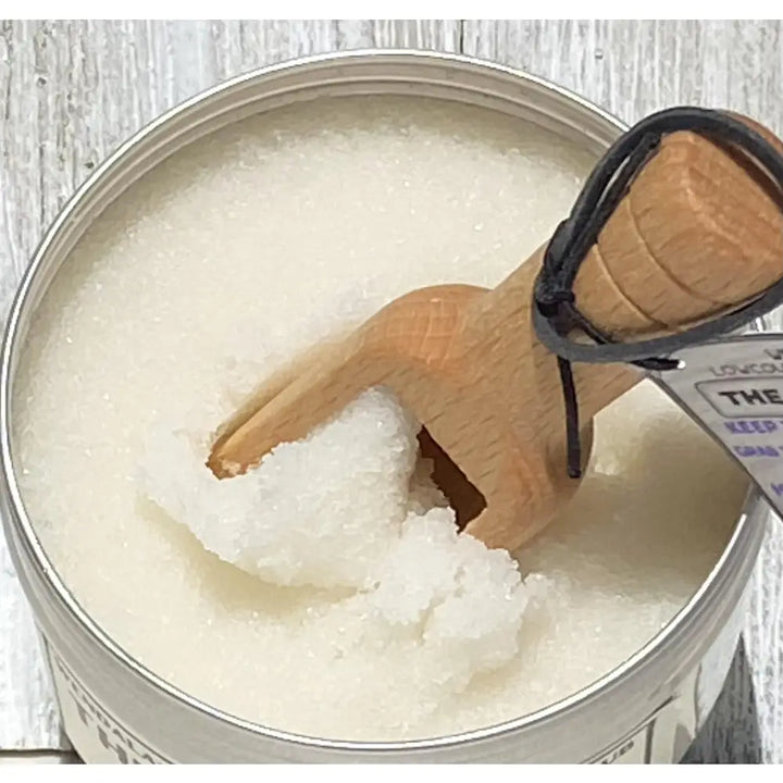 A wooden spoon with black cord handle rests in a silver tin of wendalas lemon clove organic coconut sugar hand scrub